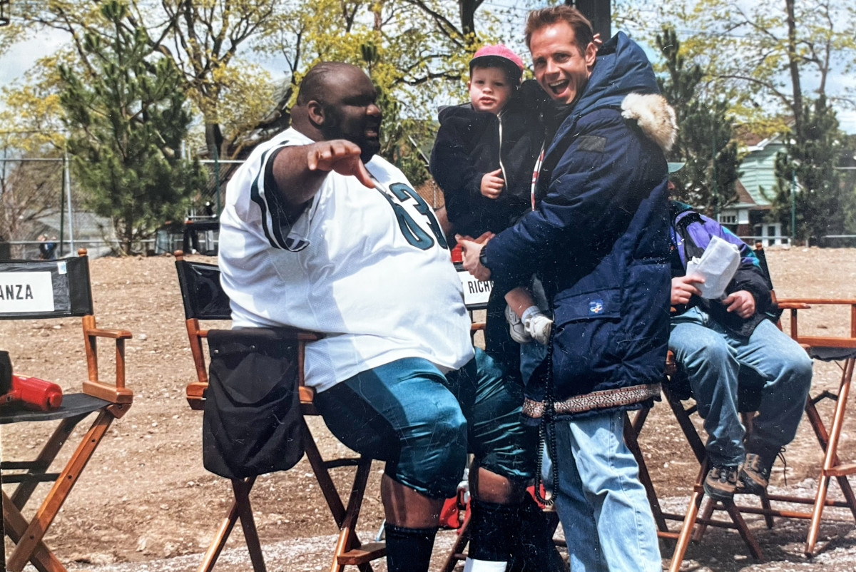 Actor Jaime Cardriche and screenwriter Tim Kelleher, holding his son, talk on set of “The Garbage Picking Field Goal Kicking Philadelphia Phenomenon”
